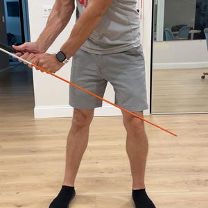 May include: A grey short-sleeved t-shirt with red and white text reading EADS, grey shorts, and black socks. The garment is worn while holding a long, thin, orange training rod with a black handle, used for practicing a golf swing motion.