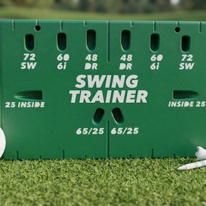 May include: A green rectangular golf swing trainer aid featuring white text that reads SWING TRAINER, 72 SW, 60 6i, 48 DR, 25 INSIDE, and 65/25. A white golf ball sits to the left, and a white golf tee is positioned to the right of the device.