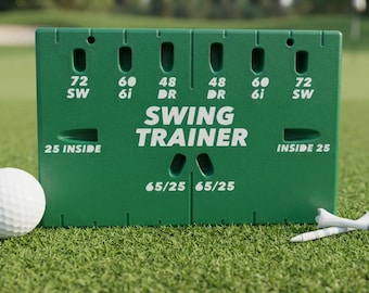 May include: A green rectangular golf swing trainer aid featuring white text that reads SWING TRAINER, 72 SW, 60 6i, 48 DR, 25 INSIDE, and 65/25. A white golf ball sits to the left, and a white golf tee is positioned to the right of the device.