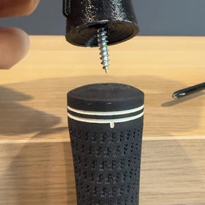May include: A black golf club grip with a white stripe detail is shown with a black screw-in end cap. The grip has a textured surface. A screw is partially inserted into the end cap.