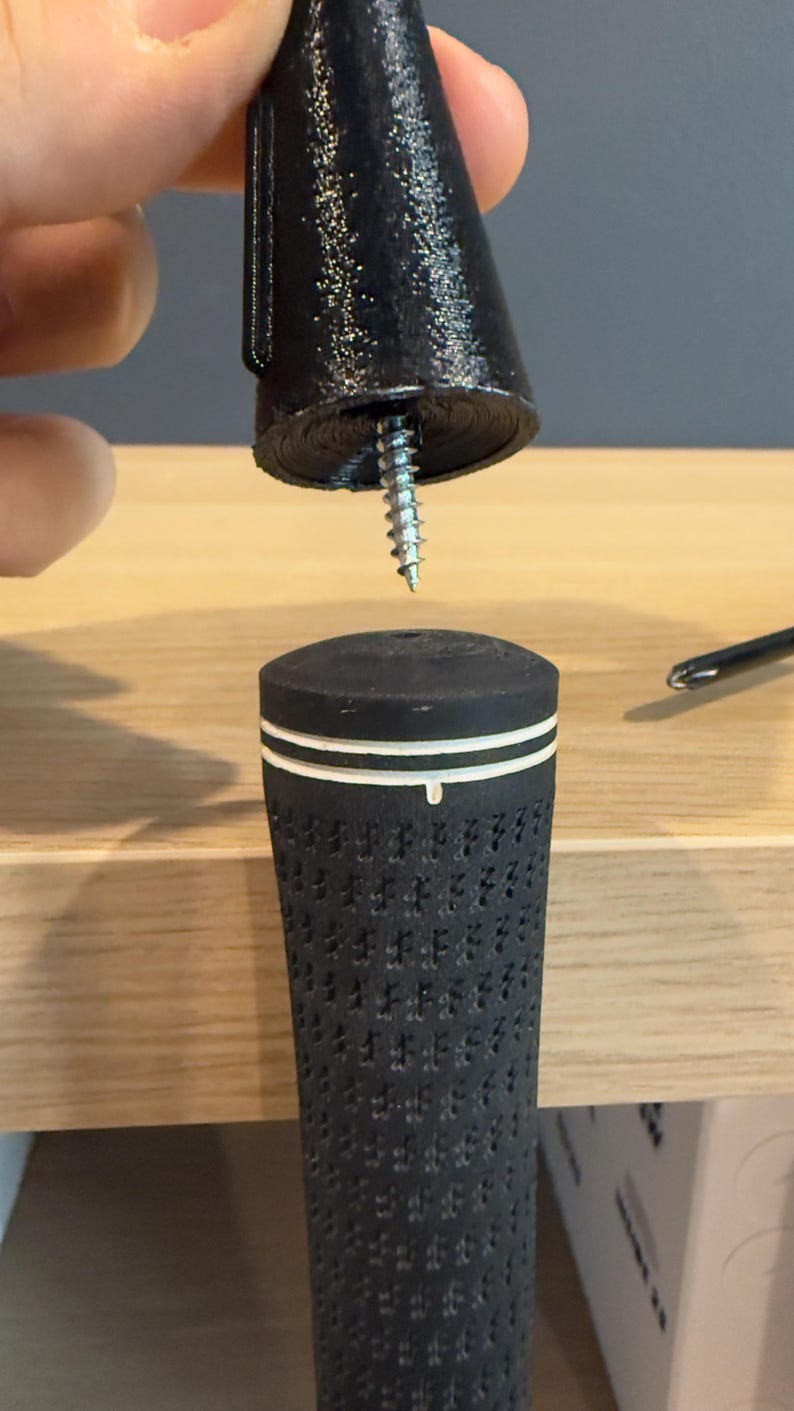 May include: A black golf club grip with a white stripe detail is shown with a black screw-in end cap. The grip has a textured surface. A screw is partially inserted into the end cap.