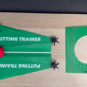 Peut inclure: Un appareil d'entraînement au putting en plastique vert avec le texte blanc PUTTING TRAINER, une balle rouge, des lignes d'alignement blanches et quatre boutons noirs réglables. À côté se trouve une coupelle de putting circulaire verte.