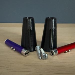 May include: An assortment of hardware components, including two black, cone-shaped pieces, two small cylindrical tools in purple and red, and several screws. The items are arranged on a light-colored wooden surface.