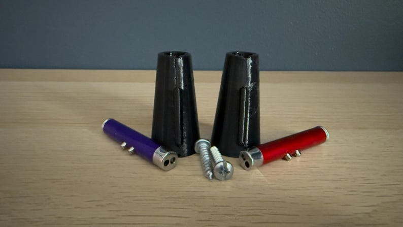 May include: An assortment of hardware components, including two black, cone-shaped pieces, two small cylindrical tools in purple and red, and several screws. The items are arranged on a light-colored wooden surface.
