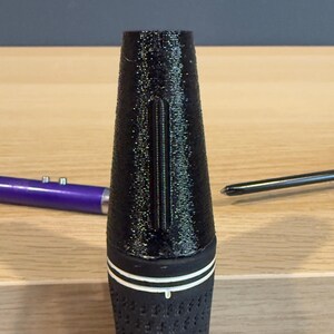 May include: Black golf club grip with a textured surface and a white stripe. The grip has a conical shape and a vertical groove. A purple pen and a screwdriver are in the background.