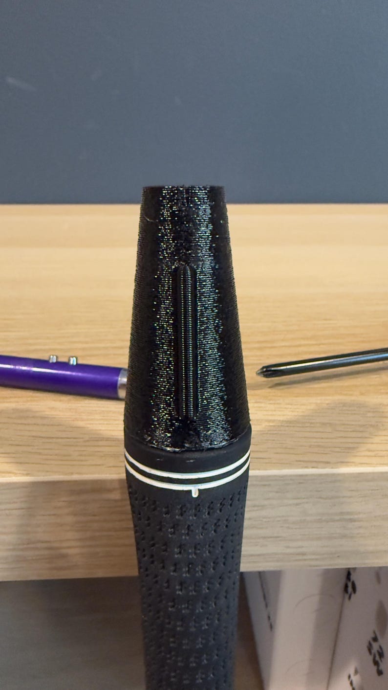 May include: Black golf club grip with a textured surface and a white stripe. The grip has a conical shape and a vertical groove. A purple pen and a screwdriver are in the background.
