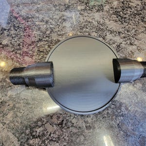 May include: A grey, circular component with two cylindrical threaded extensions. The object, likely 3D-printed, shows layer lines. The design suggests a mechanical or industrial part, possibly for plumbing or machinery.
