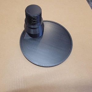 May include: A black 3D-printed object with a threaded cylindrical part on a circular base. The base has a raised edge. The object is made of a matte black material and sits on a light brown surface.
