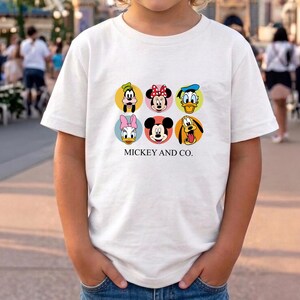 May include: A white short-sleeved t-shirt featuring a graphic print of six Disney character faces arranged in two rows of three circles. The characters include Goofy, Minnie Mouse, Donald Duck, Daisy Duck, Mickey Mouse, and Pluto. The text MICKEY AND CO. is printed in black below the circles.