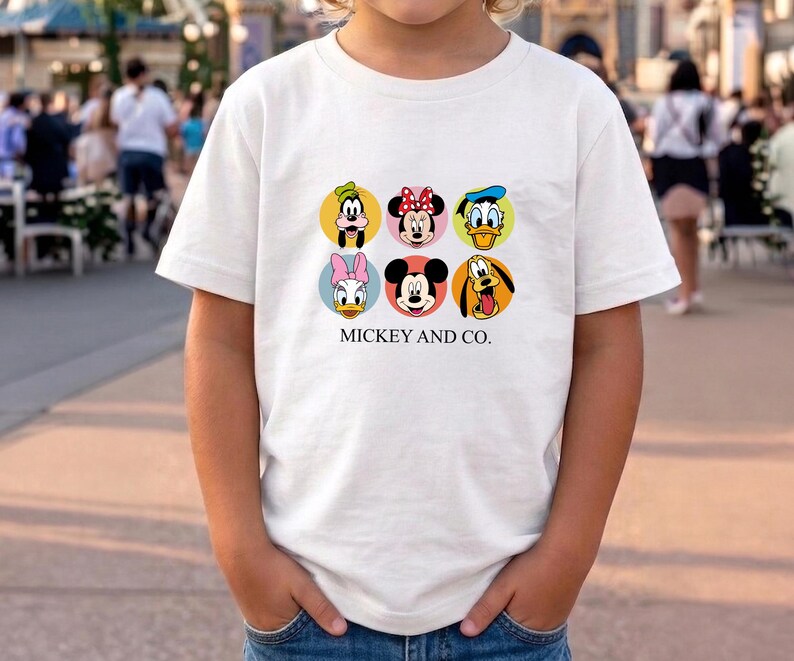 May include: A white short-sleeved t-shirt featuring a graphic print of six Disney character faces arranged in two rows of three circles. The characters include Goofy, Minnie Mouse, Donald Duck, Daisy Duck, Mickey Mouse, and Pluto. The text MICKEY AND CO. is printed in black below the circles.