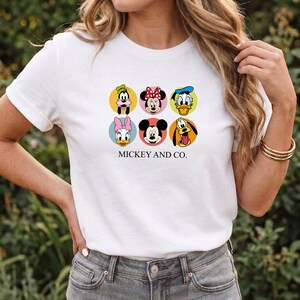 May include: A white short-sleeved t-shirt featuring a graphic print of six Disney character faces arranged in a grid within colored circles. The characters include Goofy, Minnie Mouse, Donald Duck, Daisy Duck, Mickey Mouse, and Pluto. The text MICKEY AND CO. is printed in black below the graphic.