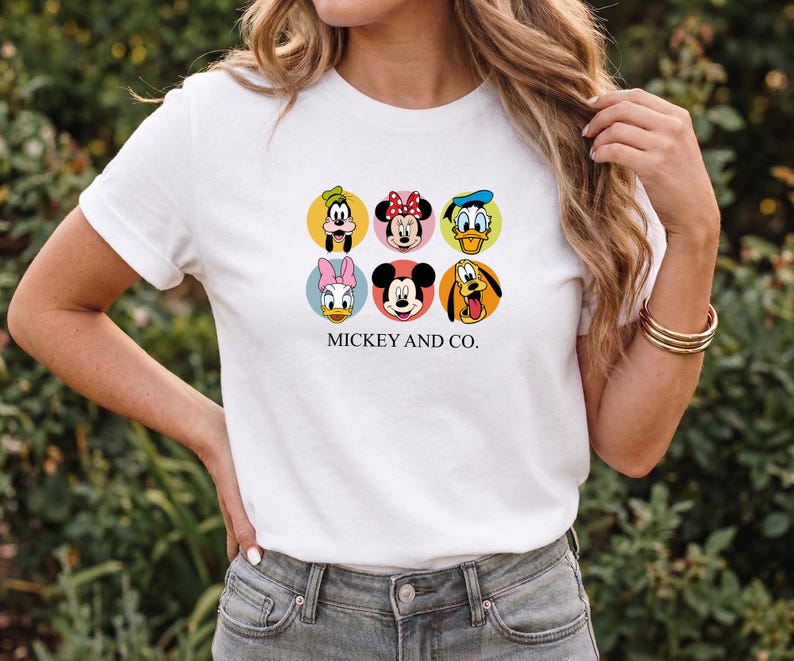 May include: A white short-sleeved t-shirt featuring a graphic print of six Disney character faces arranged in a grid within colored circles. The characters include Goofy, Minnie Mouse, Donald Duck, Daisy Duck, Mickey Mouse, and Pluto. The text MICKEY AND CO. is printed in black below the graphic.
