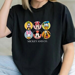 May include: A black short-sleeved t-shirt featuring a graphic print of six Disney characters in circular frames, including Goofy, Minnie Mouse, Donald Duck, Daisy Duck, Mickey Mouse, and Pluto, with the text MICKEY AND CO. printed below.