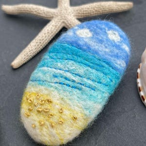 Pode incluir: Uma barra de sabão artesanal feltrada que retrata uma cena de praia. A barra apresenta um céu azul com nuvens brancas, água turquesa e uma praia de areia amarela com enfeites dourados. Uma estrela do mar está ao fundo.