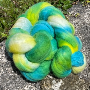 May include: A thick, soft hank of hand-dyed roving fibre featuring a vibrant gradient of bright yellow, lime green, emerald green, and turquoise blue. A light green paper label is wrapped around one section of the twisted, fluffy wool fibre.