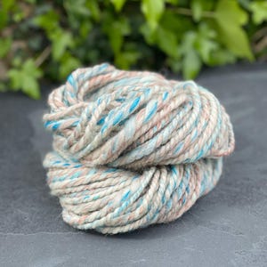 May include: A skein of yarn in a light blue, beige, and pink color combination. The yarn is twisted and has a soft, fuzzy texture.