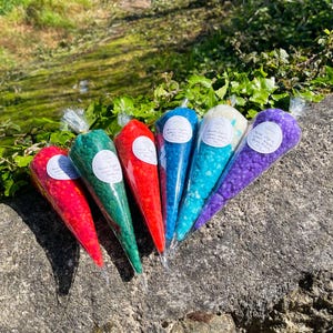May include: Six colorful cones filled with bath salts. The cones are labeled "Hand Made Soap & Bath Salts" with different scents. The cones are arranged in a row on a gray stone surface.