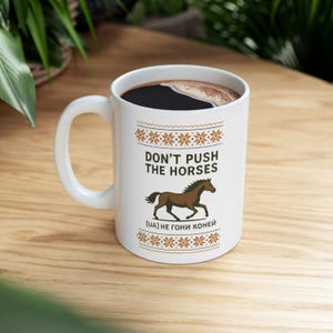 May include: White ceramic mug with dark liquid, displaying the text "DON'T PUSH THE HORSES" above a brown horse illustration. The mug has a snowflake pattern and a handle.