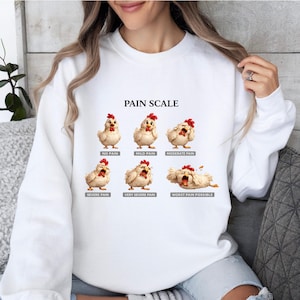 May include: White sweatshirt featuring a humorous "Pain Scale" graphic with cartoon chickens. The scale ranges from "No Pain" to "Worst Pain Possible." The text and illustrations are in black and red.