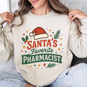 May include: Cream-colored sweatshirt with a festive design. The graphic features a Santa hat, the text "SANTA'S Favorite PHARMACIST", and decorative elements like stars and holly. The design is in red, green, and gold.
