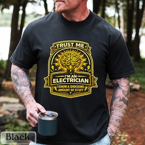 May include: Black t-shirt with a gold and yellow graphic. The graphic reads "TRUST ME" above a brain-shaped lightbulb design, with the text "I'M AN ELECTRICIAN" and "I KNOW A SHOCKING AMOUNT OF STUFF" below. A black mug is held in the hand.