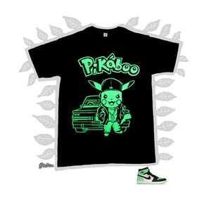 Puede incluir: Camiseta negra con un diseño verde neón de Pikachu con gorra y chaqueta, con el texto "Pi-Káboo". Se muestra una zapatilla deportiva a juego en verde neón y negro debajo.