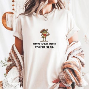 Puede incluir: Camiseta color crema con una rana de dibujos animados con sombrero de vaquero y taza de café. El texto "I HAVE TO SAY WEIRD STUFF OR I'LL DIE" está impreso debajo de la rana. La camiseta tiene mangas cortas.