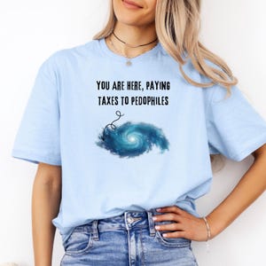 Może przedstawiać: Jasnoniebieska koszulka z napisem "YOU ARE HERE, PAYING TAXES TO PEDOPHILES" nad niebiesko-białą grafiką galaktyki. Koszulka wykonana jest z miękkiego materiału.