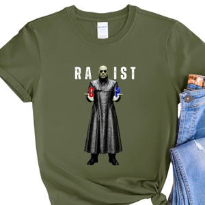 Puede incluir: Una camiseta de color verde oliva con un estampado gráfico de una persona con un abrigo largo negro y gafas de sol. La persona sostiene una letra C roja en una mano y una letra P azul en la otra. El texto blanco RA IST está impreso sobre la figura.