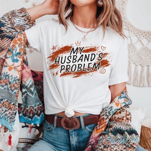 May include: White t-shirt with the text "MY HUSBAND'S PROBLEM" in a distressed font, accented with a crown, heart, star, and smiley face. The shirt is knotted at the waist and worn with blue jeans and a brown belt.