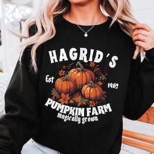 May include: Black sweatshirt featuring a graphic of three orange pumpkins with green stems and leaves. The text "Hagrid's Pumpkin Farm" is printed above the pumpkins, with "Est 1962" above the pumpkins. The phrase "magically grown" is printed below the pumpkins.