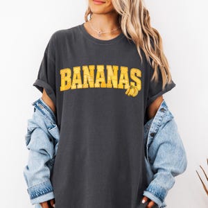 May include: A charcoal grey short-sleeve t-shirt featuring the word BANANAS in large, yellow, block-style letters with a subtle checkered pattern. A small graphic of three yellow bananas is positioned to the right of the text. The shirt is worn under a light blue denim jacket.