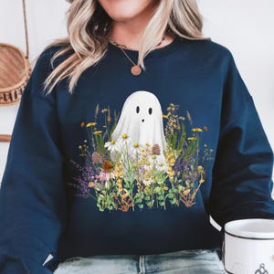 Puede incluir: Sudadera azul marino con un diseño de fantasma blanco rodeado de flores silvestres. El fantasma tiene dos ojos ovalados negros y una boca pequeña. Las flores silvestres incluyen margaritas, flores amarillas y lavanda morada.