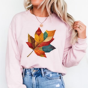 May include: A pale pink sweatshirt with a large, colourful leaf motif. The leaf is made up of various fabric patches in shades of red, orange, yellow, green, and blue, with a stitched outline. The sweatshirt is a casual, comfortable style.