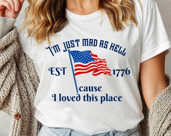 May include: White t-shirt with the text "I'M JUST MAD AS HELL" in blue, along with an American flag graphic. The shirt also includes the text "EST 1776" and "cause I loved this place" in blue.