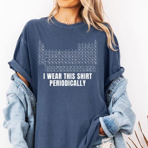 Puede incluir: Camiseta azul marino con una tabla periódica de elementos en blanco. El texto "I WEAR THIS SHIRT PERIODICALLY" está impreso debajo. La camiseta tiene mangas enrolladas y un corte holgado.