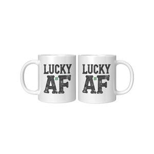 Könnte beinhalten: Zwei weiße Keramikbecher mit schwarzem Text, der "Lucky AF" lautet, und einem kleinen grünen Kleeblatt.
