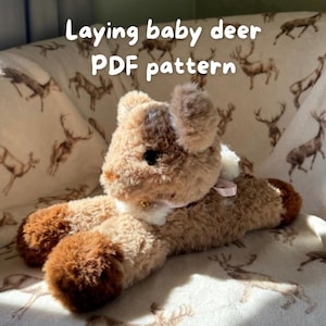 May include: A soft, laying baby deer toy in shades of brown and beige. It features a small pink ribbon and the text "Laying baby deer PDF pattern". The toy is designed to be a pattern.