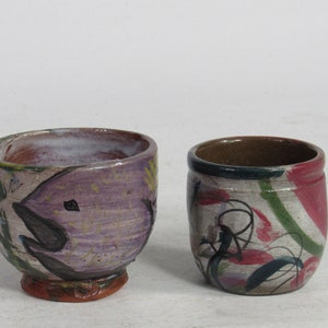 May include: Two ceramic cups with hand-painted designs. One cup is purple with a black fish design and the other is white with green, red, and black abstract designs.