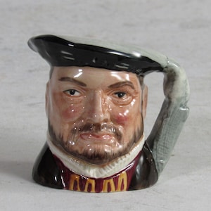 May include: A ceramic mug shaped like a man's head, featuring a black hat and a beard. The mug has a handle that extends from the hat. The man's face is detailed with painted features and a stern expression. The mug has a dark brown coat with a red and gold collar.