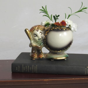 May include: A ceramic planter shaped like a baseball with a boy holding a bat. The planter is decorated with gold trim and has a brown and white glaze. The planter is filled with white and orange flowers.