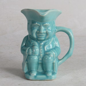 May include: A light blue ceramic pitcher shaped like a seated man wearing a hat. The pitcher has a handle and a spout.