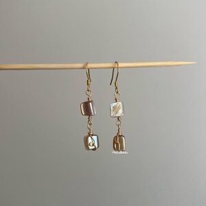 May include: A pair of drop earrings featuring two square-shaped, iridescent shell beads suspended from gold-toned wire hooks. Each bead is wrapped in thin gold-toned wire, creating a dangling design that hangs from a horizontal wooden stick.