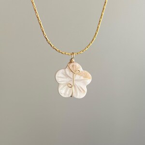 Op de afbeelding: Een goudkleurige ketting met een bloemhanger. De hanger is wit met een gesneden bloemmotief en een goudkleurige draadwikkeling. De ketting wordt getoond tegen een neutrale achtergrond.