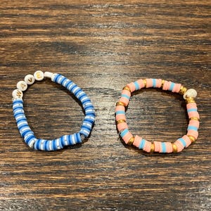 以下が含まれることがあります： 2つのビーズブレスレット。1つのブレスレットは、青、白、水色のビーズが特徴で、白い文字ビーズで「WAVE」という単語が綴られています。もう1つのブレスレットは、ピンク、水色、ゴールドのビーズが付いています。