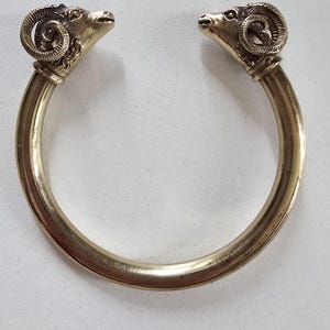 Peut inclure: Un bracelet manchette en métal doré orné de deux têtes de bélier détaillées se faisant face aux extrémités ouvertes. L'anneau est lisse et circulaire, tandis que les têtes de bélier présentent des cornes texturées et des traits faciaux.