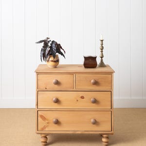 May include: A light brown wooden chest of drawers with two small top drawers and two larger bottom drawers, each featuring round wooden knobs. The piece stands on four turned feet and is topped with a potted plant, a small dark box, and a candle holder.