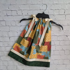 May include: A patchwork dress with a green, blue, yellow, and orange floral print. The dress has a white ribbon tied at the shoulders and a green trim around the bottom.