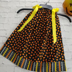 K&ouml;nnte beinhalten: Ein schwarzes, &auml;rmelloses Kleid mit einem Candy-Corn-Druck und gelben Schleifenb&auml;ndern. Das Kleid hat einen schwarz-wei&szlig;en Streifenbesatz mit orangefarbenen und gelben Akzenten.