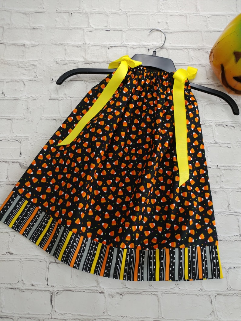 K&ouml;nnte beinhalten: Ein schwarzes, &auml;rmelloses Kleid mit einem Candy-Corn-Druck und gelben Schleifenb&auml;ndern. Das Kleid hat einen schwarz-wei&szlig;en Streifenbesatz mit orangefarbenen und gelben Akzenten.